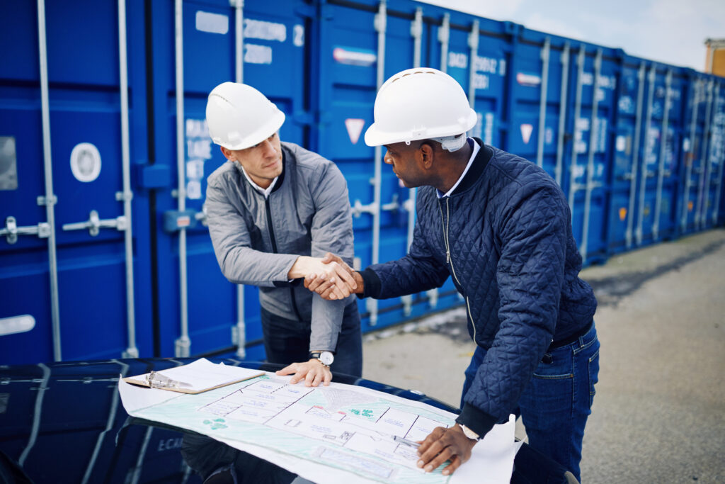Two engineers in hard hats shaking hands over drawings in a freight yard